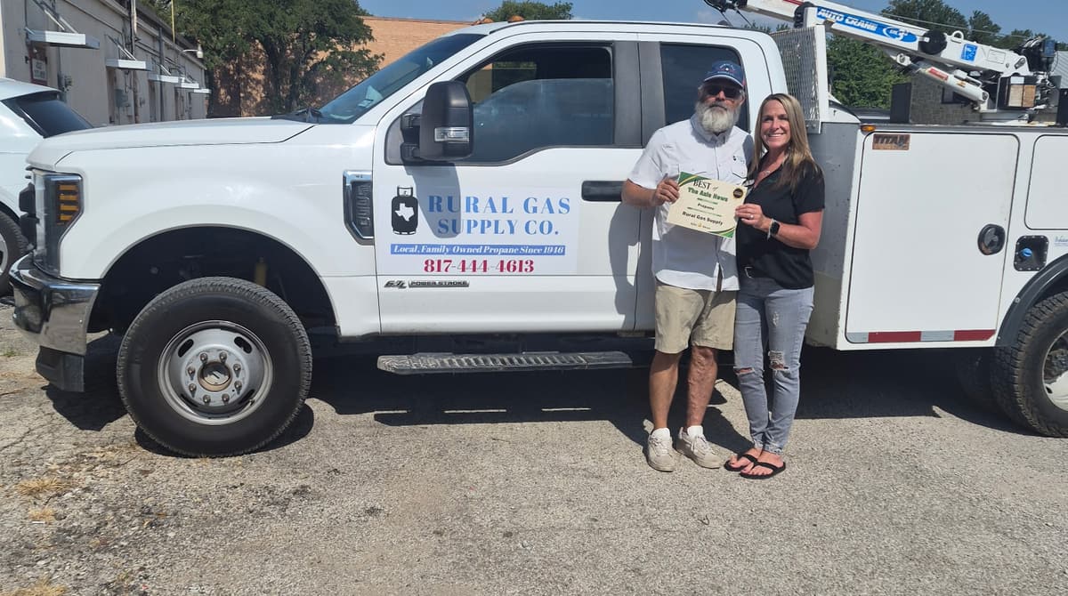 Two Rural Gas Supply team members stand beside a company service truck, one holding a printed certificate.