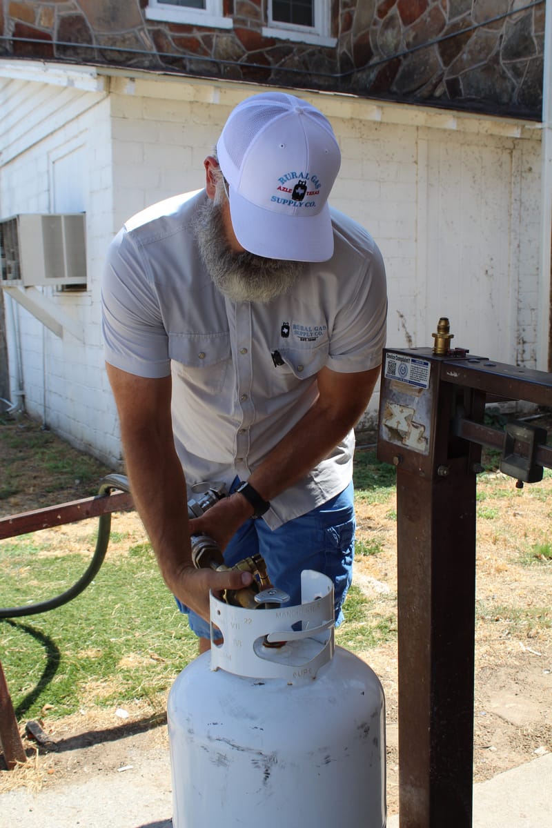 A Rural Gas Supply crew member filling a portable propane cylinder at the yard fill stand, the shop building behind.